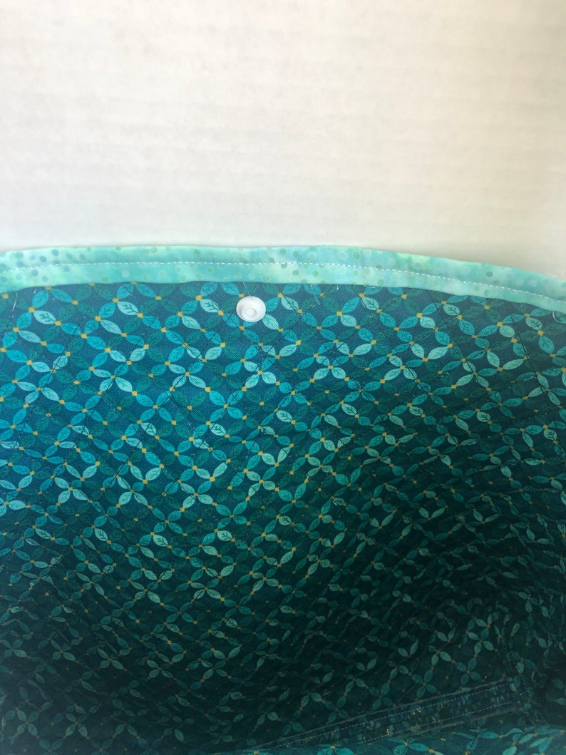 May include: The inside of a teal fabric bag with a white button closure. The fabric has a pattern of small teal leaves on a dark teal background.