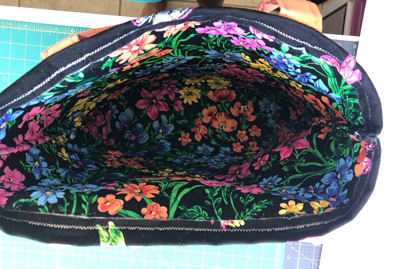 May include: A black tote bag with a zipper closure. The inside of the bag is lined with a colorful floral fabric.