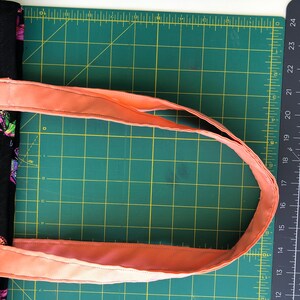 May include: A close-up of a peach-colored fabric strap for a bag, laid out on a green cutting mat. The strap is folded in half and is ready to be sewn.