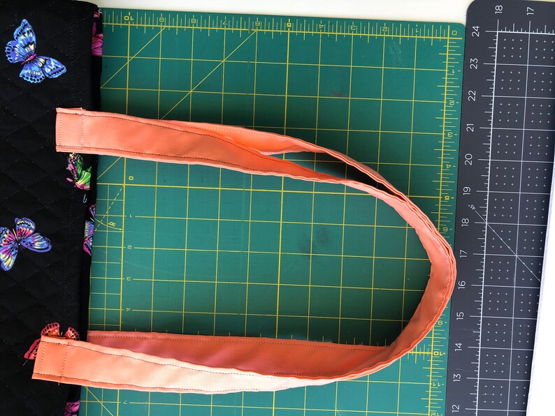 May include: A close-up of a peach-colored fabric strap for a bag, laid out on a green cutting mat. The strap is folded in half and is ready to be sewn.