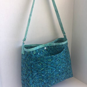 May include: A teal blue fabric tote bag with a green and blue floral pattern. The bag has a single pocket on the front and a long teal blue strap.