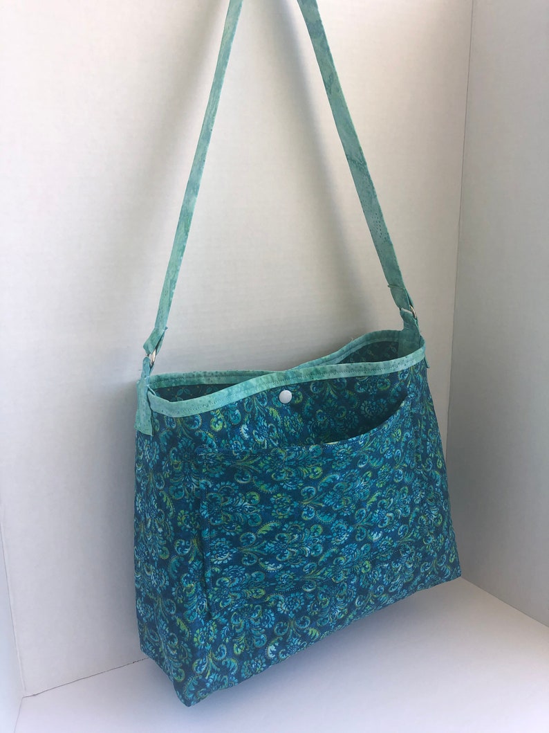 May include: A teal blue fabric tote bag with a green and blue floral pattern. The bag has a single pocket on the front and a long teal blue strap.
