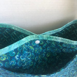 May include: A teal blue fabric tote bag with a white button closure. The bag has a floral pattern with gold accents.