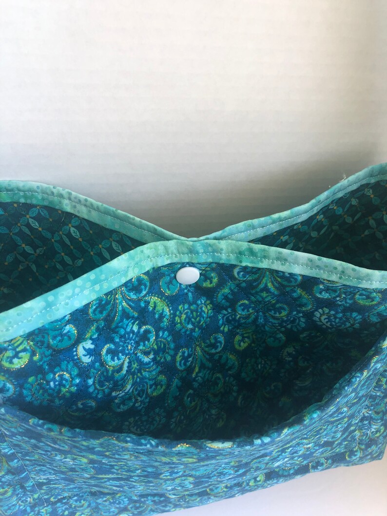 May include: A teal blue fabric tote bag with a white button closure. The bag has a floral pattern with gold accents.