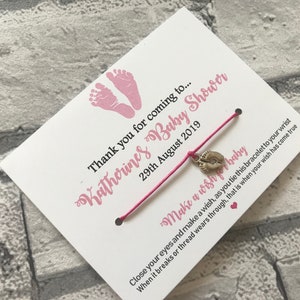 May include: A pink and white card with the text "Katherines Baby Shower" and "Thank you for coming to... 29th August 2019". A pink string bracelet with a silver charm of a baby footprint is attached to the card. The text "Make a wish for baby" is written on the card. The text "Close your eyes and make a wish, as you tie this bracelet to your wrist. When it breaks or thread wears through, that is when your wish has come true." is also written on the card.