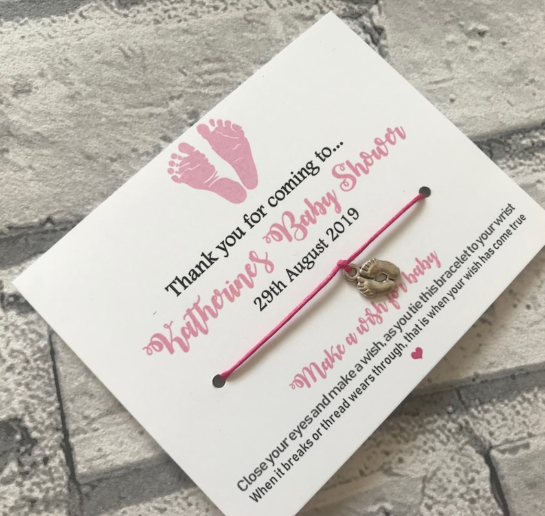 May include: A pink and white card with the text "Katherines Baby Shower" and "Thank you for coming to... 29th August 2019". A pink string bracelet with a silver charm of a baby footprint is attached to the card. The text "Make a wish for baby" is written on the card. The text "Close your eyes and make a wish, as you tie this bracelet to your wrist. When it breaks or thread wears through, that is when your wish has come true." is also written on the card.