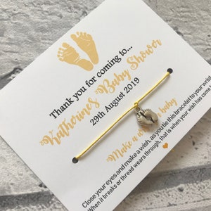 May include: A yellow string bracelet with a silver charm of two baby footprints. The bracelet is attached to a white card with the text "Thank you for coming to... Katherines Baby Shower 29th August 2019" and "Make a wish... baby Close your eyes and make a wish, as you tie this bracelet to your wrist When it breaks or thread wears through, that is when your wish has come true".