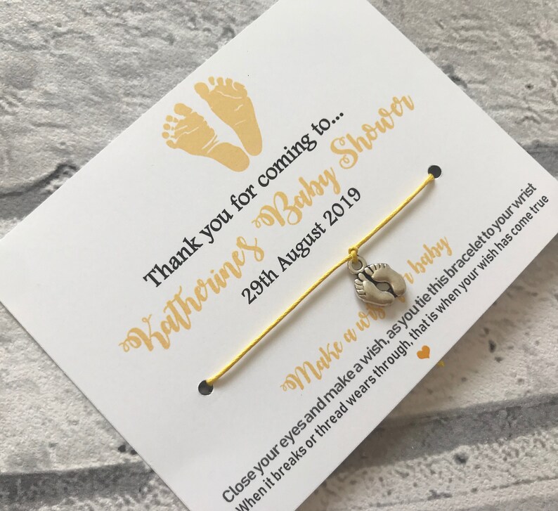 May include: A yellow string bracelet with a silver charm of two baby footprints. The bracelet is attached to a white card with the text "Thank you for coming to... Katherines Baby Shower 29th August 2019" and "Make a wish... baby Close your eyes and make a wish, as you tie this bracelet to your wrist When it breaks or thread wears through, that is when your wish has come true".