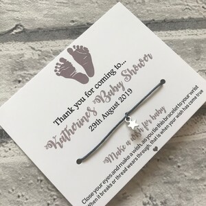 May include: A white card with grey baby footprints and the text "Katherines Baby Shower" and "Thank you for coming to... 29th August 2019". A grey string bracelet with a silver star charm is attached to the card. The card reads "Make a wish for baby Close your eyes and make a wish, as you tie this bracelet to your wrist When it breaks or thread wears through, that is when your wish has come true."