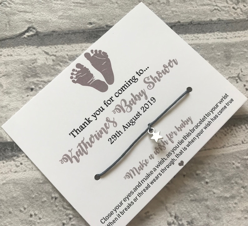 May include: A white card with grey baby footprints and the text "Katherines Baby Shower" and "Thank you for coming to... 29th August 2019". A grey string bracelet with a silver star charm is attached to the card. The card reads "Make a wish for baby Close your eyes and make a wish, as you tie this bracelet to your wrist When it breaks or thread wears through, that is when your wish has come true."