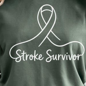 May include: A dark olive green short-sleeved t-shirt featuring a white line-art graphic of a ribbon centred on the chest. Below the ribbon, the text "Stroke Survivor" is printed in a white, cursive, script-style font across the front.