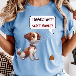 May include: Light blue t-shirt featuring a cartoon dog with brown and white fur, a speech bubble with the text "I SAID SIT! NOT S#%T!", and a small pile of poop. The dog is wearing a collar.