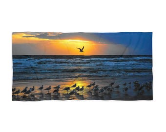 May include: Beach towel depicting a sunset over the sea. A flock of seagulls stands on the wet sand, with one bird in flight. The sky is a blend of orange, yellow, and blue, with the sun's rays breaking through the clouds.