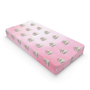 Könnte beinhalten: Ein rosa Wickeltischbezug mit einem sich wiederholenden Muster eines schlafenden Babys und dem Text "Angel Baby". Der Bezug hat einen Farbverlauf von hellrosa zu dunklerem Rosa.