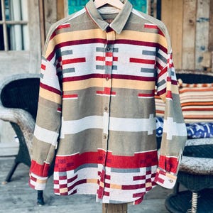 May include: A long-sleeved shirt with a geometric pattern in red, yellow, grey, and white. The shirt is hanging on a wooden hanger. It has a collared neckline and buttoned cuffs. The shirt's design is made up of horizontal and vertical lines.