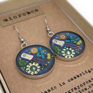 May include: A pair of round earrings with a microbe design. Each earring has a silver-coloured frame and a dark blue interior filled with colourful, miniature microbe shapes. The earrings are displayed in a brown box with the word "microbes" printed on a small tag.