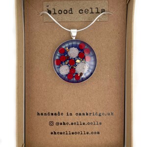Puede incluir: Un collar con colgante redondo, con un fondo azul con dise&ntilde;os circulares rojos y blancos, y motas doradas. El colgante se muestra en una caja de cart&oacute;n marr&oacute;n con el texto "blood cells".