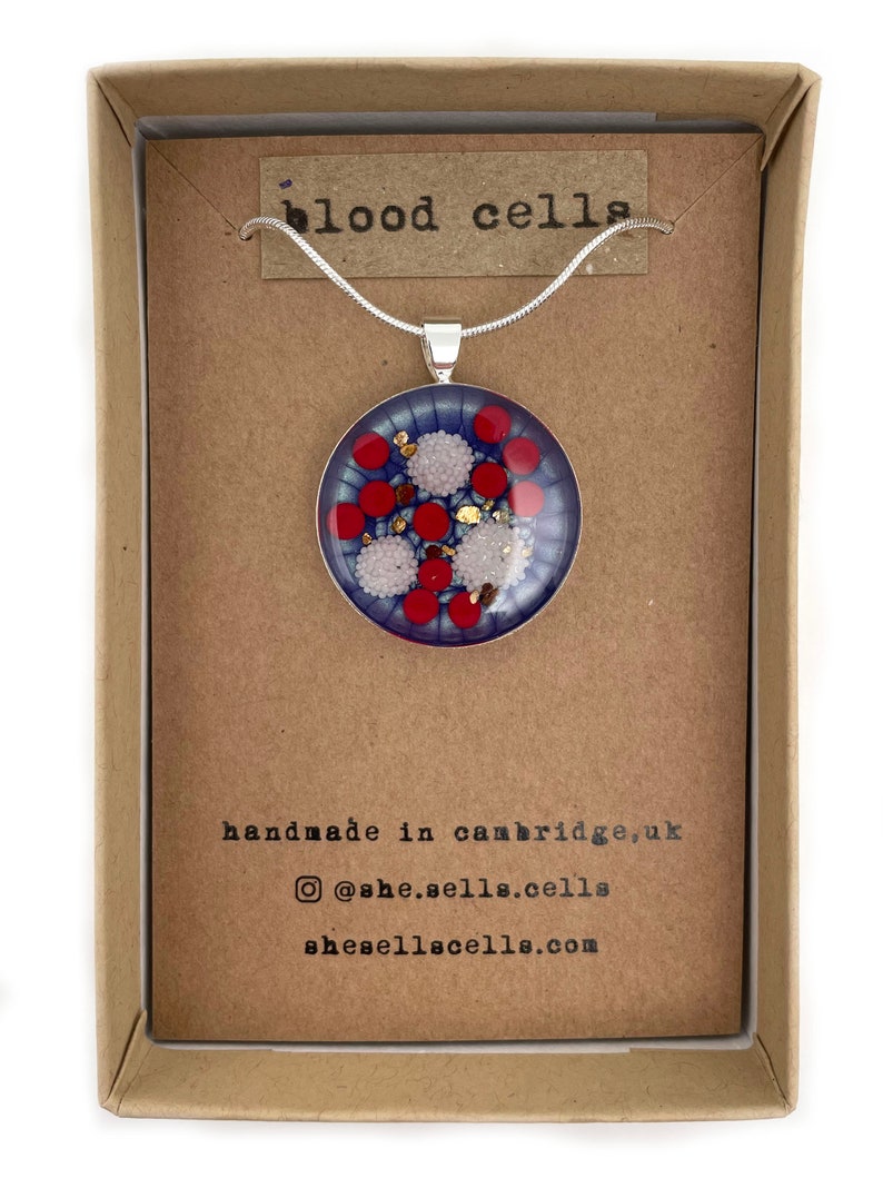 Puede incluir: Un collar con colgante redondo, con un fondo azul con dise&ntilde;os circulares rojos y blancos, y motas doradas. El colgante se muestra en una caja de cart&oacute;n marr&oacute;n con el texto "blood cells".