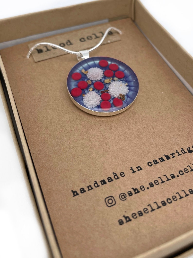 Puede incluir: Un collar con colgante de plata con un dise&ntilde;o circular. El colgante presenta elementos rojos, blancos y dorados, que se asemejan a c&eacute;lulas sangu&iacute;neas, sobre un fondo azul. Presentado en una caja de cart&oacute;n marr&oacute;n. El texto "blood cells" es visible.