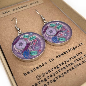 Puede incluir: Un par de pendientes de plata con un diseño de célula animal morado y verde. Los pendientes están hechos a mano en Cambridge, Reino Unido. El texto "the animal cell" está en la caja.