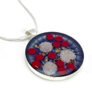 Puede incluir: Un collar con colgante plateado con un colgante redondo de tono azulado. El colgante presenta elementos circulares rojos y blancos, junto con peque&ntilde;os acentos dorados. El collar tiene una cadena plateada.