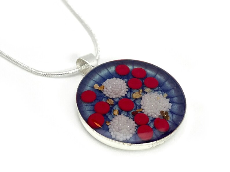 Puede incluir: Un collar con colgante plateado con un colgante redondo de tono azulado. El colgante presenta elementos circulares rojos y blancos, junto con peque&ntilde;os acentos dorados. El collar tiene una cadena plateada.