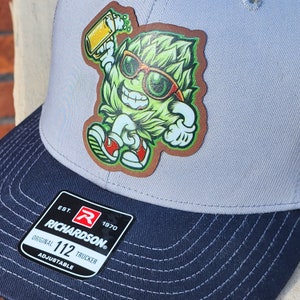 May include: A gray and navy blue trucker hat with an adjustable strap. The hat features a green, cartoon character wearing sunglasses and holding a glass of beer. The text "EST. 1970 RICHARDSON. ORIGINAL 112 TRUCKER ADJUSTABLE" is printed on a white label attached to the hat.