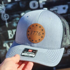 May include: A light blue trucker hat with a black mesh back. The hat features a brown leather patch with the year "1776" and star accents. The hat is adjustable and has a Richardson brand tag.