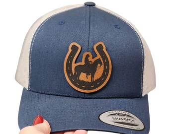 May include: A navy blue and white trucker hat with a leather patch featuring a cowboy on a horse inside a horseshoe. The hat has a curved brim and a snapback closure. The text "THE AUTHENTIC SNAPBACK" is on a tag.