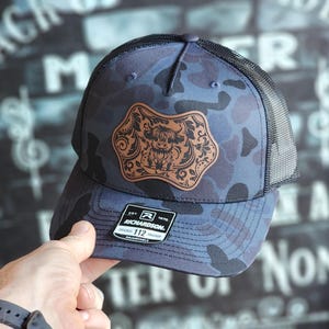 May include: A blue camouflage trucker hat with a brown leather patch featuring a Highland cow design. The hat is adjustable and has a black mesh back.