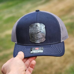 May include: Navy blue and gray trucker hat with a silver emblem that reads "BALES AND BULLIGANS". The hat has a curved brim and a mesh back. The hat is being held by a person.