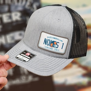 May include: Gray trucker hat with a black mesh back and a license plate-style patch that reads "NOLES 1 SUNSHINE STATE" with the website myflorida.com. The hat has a curved brim and an adjustable strap.