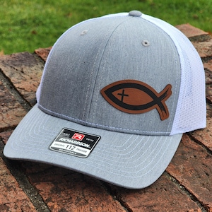 May include: A gray and white trucker hat with a brown leather fish symbol with a cross on it. The hat has an adjustable strap and a Richardson 112 label.