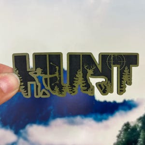 May include: A die-cut sticker featuring the word HUNT in bold, olive green lettering against a black background. The design incorporates silhouettes of a person using a bow and arrow, a deer, pine trees, and a circular crosshair target symbol.