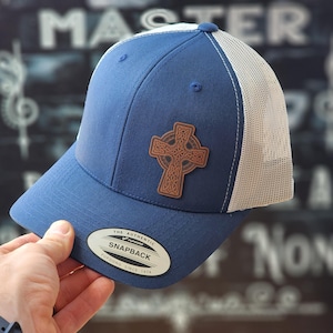 May include: A blue and white baseball cap with a brown leather Celtic cross patch. The cap is a trucker style with a mesh back.