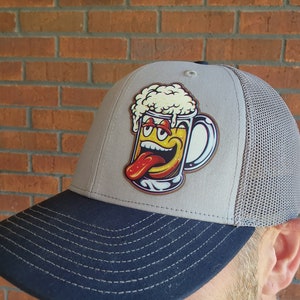 May include: A gray and black trucker hat with a cartoon image of a beer mug with a smiling face and a tongue sticking out. The mug is filled with yellow beer and white foam.
