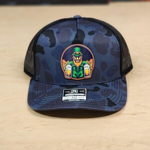 May include: A blue and black camouflage trucker hat with a leather patch featuring a cartoon leprechaun holding two beers. The patch is brown with green, yellow, and white accents. The hat has a black mesh back and a Richardson 112 label.