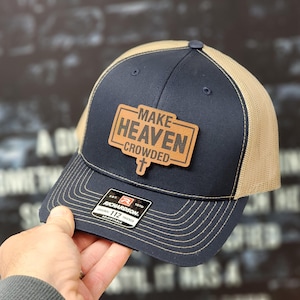 May include: A navy blue and tan trucker hat with a brown leather patch that says "Make Heaven Crowded" with a cross symbol.