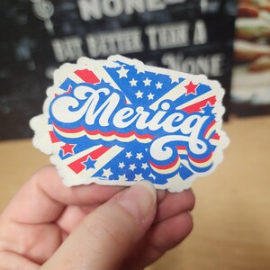 May include: A sticker with a patriotic design featuring the word "Merica" in white, with a blue background filled with white stars. Red and white stripes and accents complete the design. The sticker is held in a hand.
