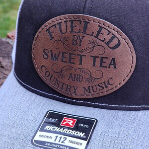 May include: A black and gray trucker hat with a brown leather patch that says "Fueled by Sweet Tea and Country Music". The hat has a white mesh back and a Richardson logo with the text "Est. 1970 Original 112 Trucker Adjustable".