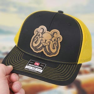 May include: Black and yellow trucker hat with a leather patch featuring a skull and octopus design. The hat has a mesh back and a curved brim. The patch is tan and the hat has the text "RICHARDSON" and "112 TRUCKER".