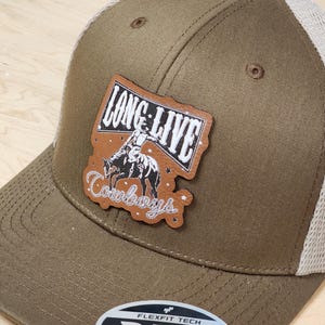 May include: A brown trucker hat with a mesh back and a curved brim. The hat features a leather patch with the words "LONG LIVE Cowboys" and an illustration of a cowboy riding a horse. The hat has a Flexfit Tech label.