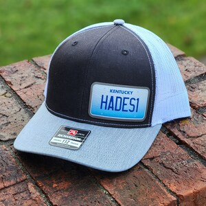 May include: A black and light blue trucker hat with a white mesh back. The hat has a patch on the front that says "Kentucky Hades1".