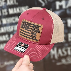 May include: A red and beige trucker hat with a brown leather patch featuring an American flag and the text "MAMA. MOMMY. MOM. BRUH."