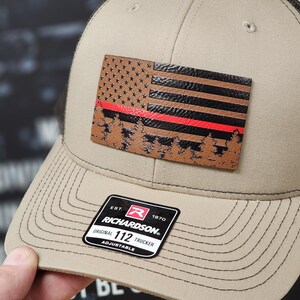 May include: A tan and black trucker hat with a brown leather patch featuring an American flag with a red stripe across the middle and a forest scene at the bottom. The patch has the text "EST. 1970 RICHARDSON. ORIGINAL 112 TRUCKER ADJUSTABLE" on the bottom.