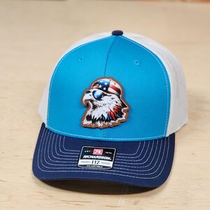 May include: A blue and white trucker hat with a brown leather patch of an eagle wearing sunglasses and a red, white, and blue bandana. The hat has a navy blue brim and a white mesh back. The hat has a Richardson 112 Original Trucker label.