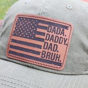 May include: Olive green baseball cap with a brown leather patch featuring an American flag design and the words "DADA. DADDY. DAD. BRUH." The cap has a curved brim and a stitched design.