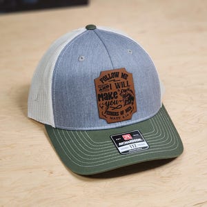 May include: A gray and green trucker hat with a brown leather patch that says "Follow me and I will make you fishers of men Matt 4:19".