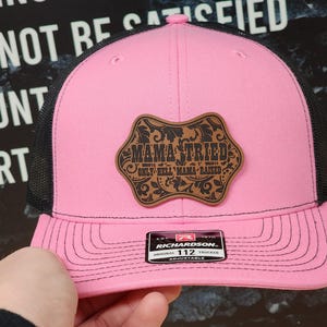 May include: Pink trucker hat with a black mesh back and a curved brim. The hat features a brown leather patch with the words "MAMA TRIED ONLY HELL MAMA RAISED" in a decorative font. The hat is adjustable.
