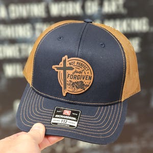May include: A navy blue and brown trucker hat with a brown leather patch that says "Not Perfect Just Forgiven" with a cross and floral design.